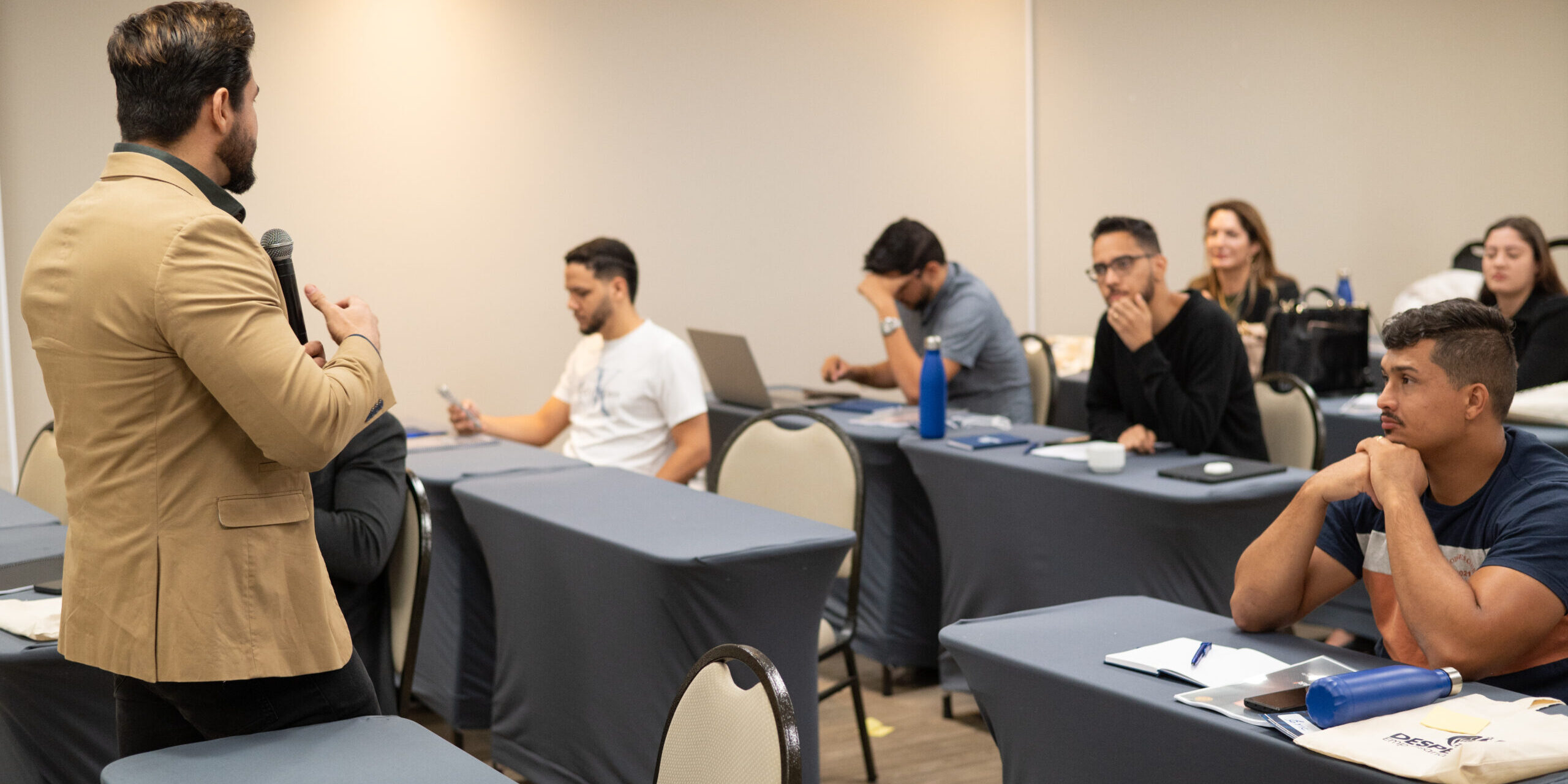 Janderson Santos conduzindo treinamento de vendas em sala com mesas e participantes Janderson Santos conduzindo treinamento de vendas em sala com mesas e participantes